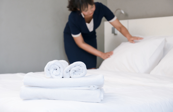 Hotel room service. Maid making bed in a room, focus of clean towels